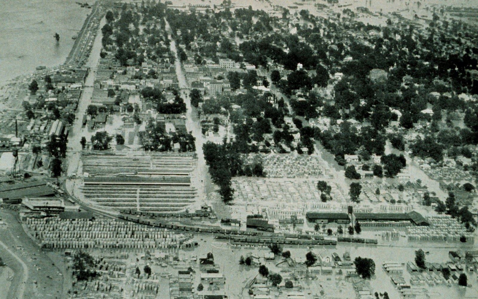 Aerial view of a flooded town with rows of storage sheds and railway lines along a riverbank