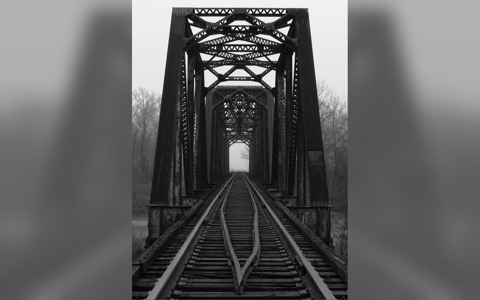 Steel truss railroad bridge with tracks receding into misty distance, bare trees on riverbank